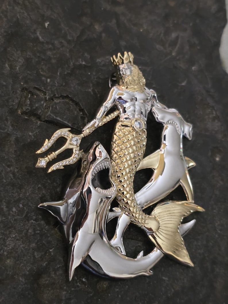 May include: A two-tone metal pendant featuring a merman with a trident, a crown, and a fish tail, flanked by two sharks. The merman is gold-colored, and the sharks are silver-colored. The pendant has a detailed design with a gemstone on the merman's chest.