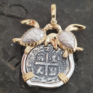 May include: A gold and silver pendant featuring a coin with a cross design, framed by a silver border and accented with gold turtle figures. The pendant has a gold bail for hanging.