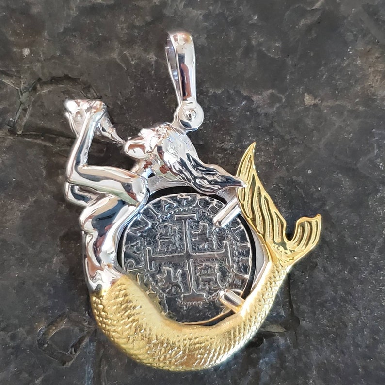 May include: A silver and gold pendant featuring a mermaid holding a coin with a cross design. The mermaid's tail is gold and the coin is silver.