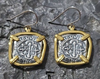 Könnte beinhalten: Ein Paar silber-goldener Ohrringe. Jeder Ohrring hat ein quadratisches, silbernes Münzdesign mit einem Kreuzmotiv, das in Gold eingefasst ist. Die Ohrringe haben einen goldenen Rand und silberne Ohrhaken. Die Ohrringe werden auf einer strukturierten, dunkelgrauen Oberfläche präsentiert.