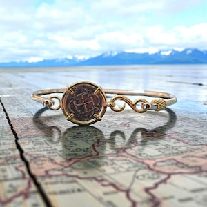 May include: A gold-toned bracelet featuring a large, round, antique-style coin at its center. The coin is set in a gold bezel and has intricate markings. The bracelet has a delicate design with decorative elements and a rope-like clasp. The background shows a map and a scenic view.