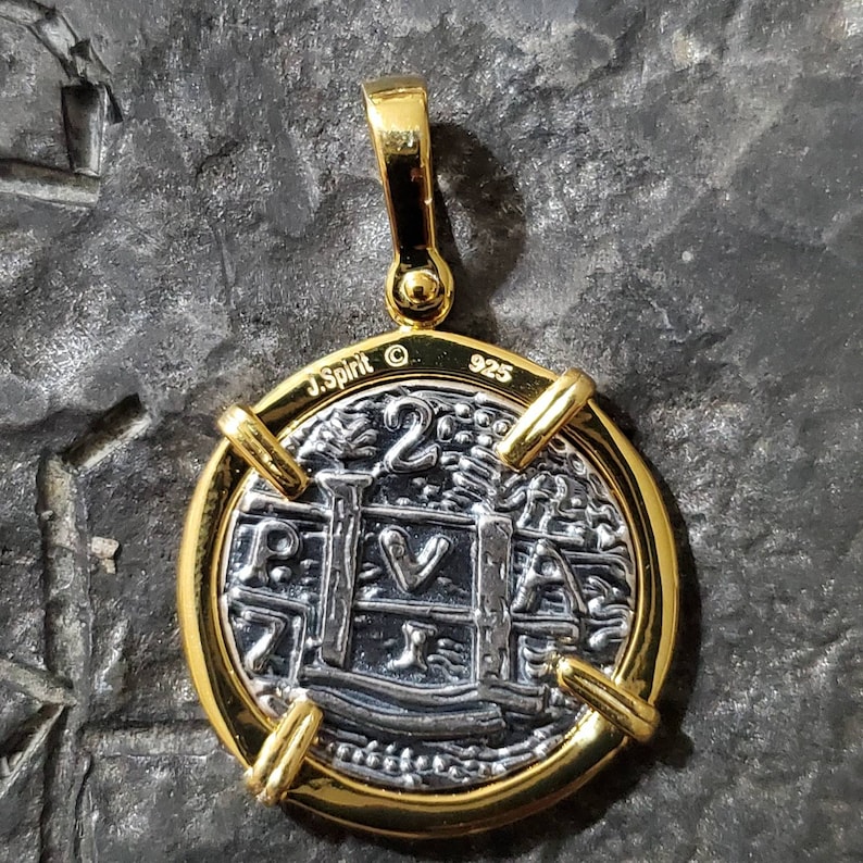 Pu&ograve; includere: Un ciondolo a forma di moneta d'argento con una cornice dorata e un'anellina dorata. La moneta presenta un disegno di un edificio con una croce in cima e il testo "2", "P", "V", "A" e "I". Il retro del ciondolo &egrave; contrassegnato con "J. Spirit &copy; 925".