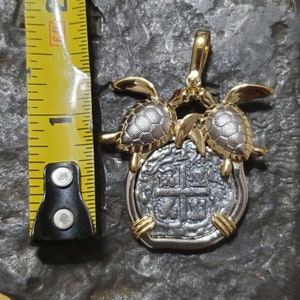 May include: A silver and gold pendant featuring a coin design with a cross and other markings. Two gold turtles are on top of the coin. The pendant is approximately 1 inch in length.