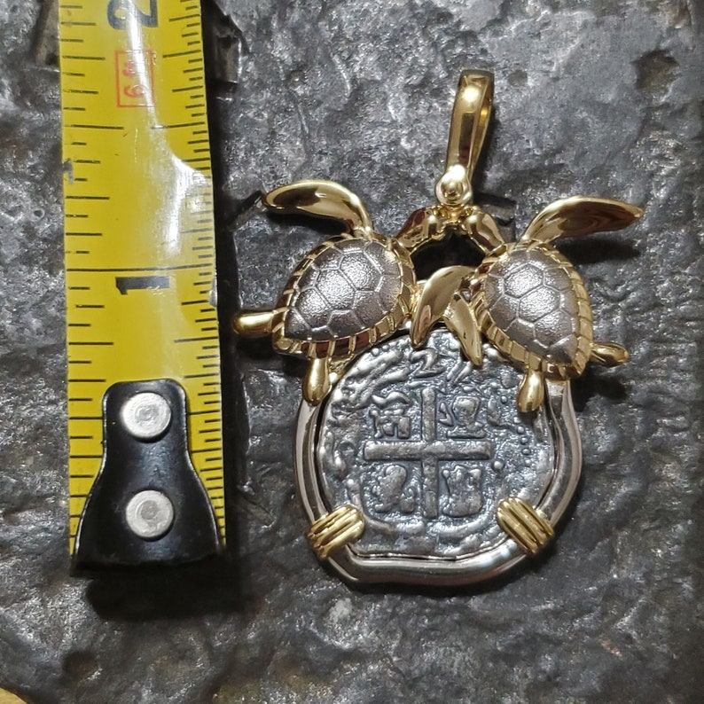 May include: A silver and gold pendant featuring a coin design with a cross and other markings. Two gold turtles are on top of the coin. The pendant is approximately 1 inch in length.