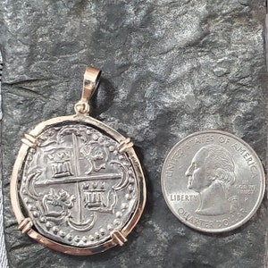 May include: A silver coin pendant with a gold frame. The coin features a cross with four shields and a crescent moon. The pendant is about the size of a US quarter.