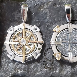 May include: Two silver and gold pendants with a compass and a coin design. The compass pendant has a gold star with a diamond in the center. The coin pendant has a silver coin with a cross design. Both pendants have a bail for attaching to a chain.