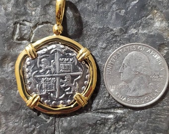 Puede incluir: Un colgante de moneda de plata con marco de oro. La moneda presenta una cruz con cuatro escudos y una corona. El colgante es aproximadamente del tamaño de una moneda de 25 centavos de dólar estadounidense.
