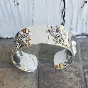 May include: A silver cuff bracelet with an octopus and a starfish design. The bracelet is open and has a smooth, polished finish.