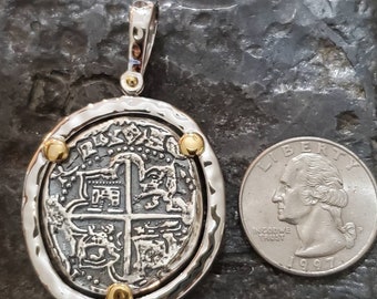 Puede incluir: Un colgante de plata y oro con una réplica de una moneda española de ocho reales. La moneda tiene un diseño detallado con una cruz y cuatro escudos. El colgante está rodeado por un bisel de plata con cuatro acentos dorados. El colgante es aproximadamente del tamaño de una moneda de euro.