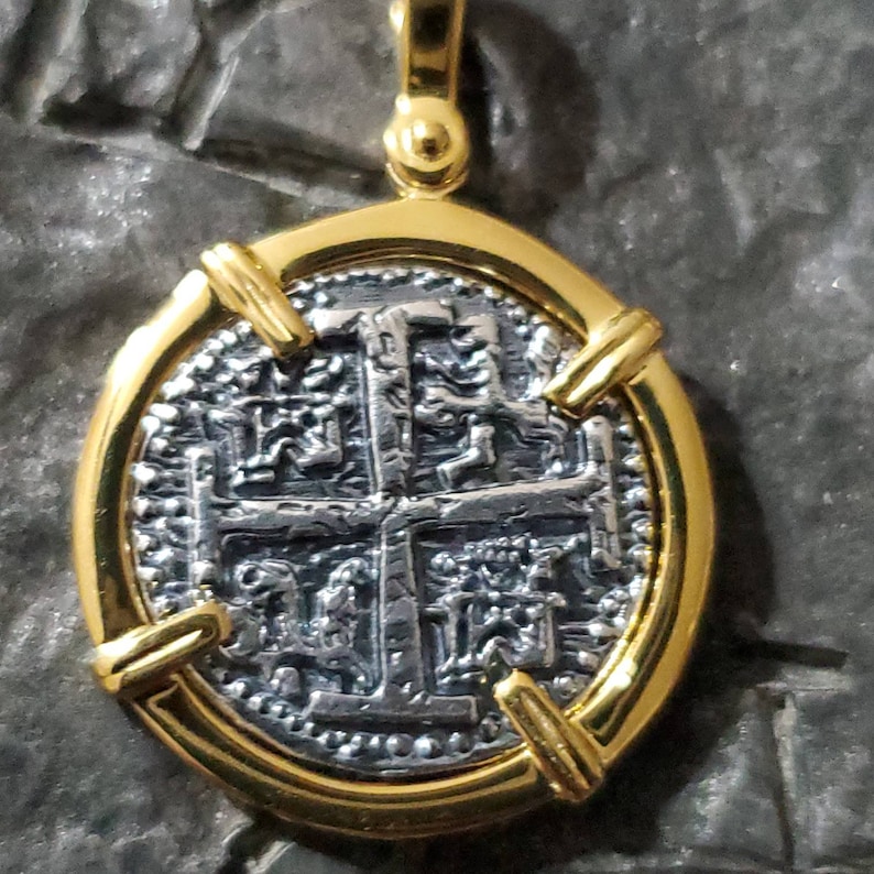 Pu&ograve; includere: Un ciondolo placcato oro con una replica in argento di una moneta spagnola da otto reali. La moneta presenta un motivo a croce con dettagli intricati ed &egrave; circondata da una cornice dorata.