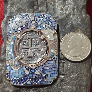 May include: A silver pendant with a blue and white mosaic design. The pendant features a silver coin with a cross and the text "ATOCHA 1622".