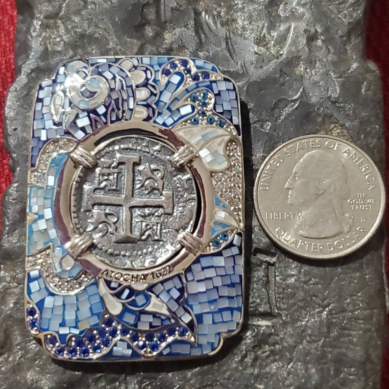 May include: A silver pendant with a blue and white mosaic design. The pendant features a silver coin with a cross and the text "ATOCHA 1622".