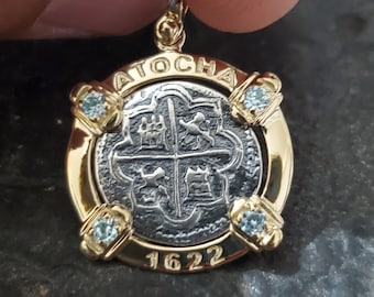 Può includere: Un ciondolo d'oro con una moneta d'argento che presenta l'iscrizione "ATOCHA 1622" e quattro stemmi. Il ciondolo è adornato con quattro pietre preziose blu.