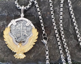 May include: A silver chain necklace with a gold eagle pendant featuring a silver coin with a cross and two figures. The coin is surrounded by a circle of clear stones.