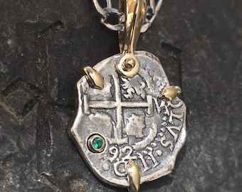 Puede incluir: Un collar con colgante de plata con un dije en forma de moneda. El dije tiene detalles dorados, una pequeña gema verde y detalles grabados. Cuelga de una cadena de plata. El colgante tiene un aspecto vintage.