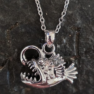 May include: A silver-colored anglerfish pendant necklace. The pendant features a detailed anglerfish design with an open mouth and sharp teeth. The necklace has a delicate chain.