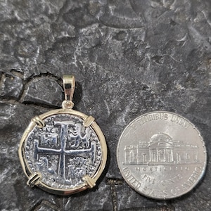 May include: A silver coin pendant with a gold-colored bezel, featuring a cross design. Next to it is a US nickel coin for size comparison. The pendant is on a dark, textured surface.