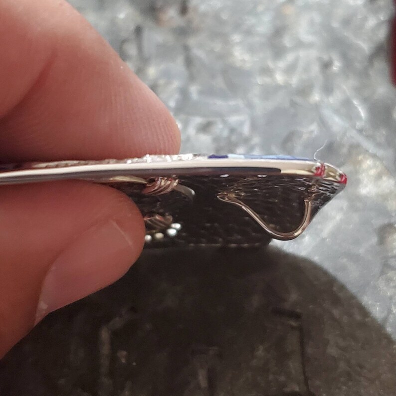 May include: A silver belt buckle with a textured surface and a blue enamel design. The buckle has two metal loops on the back for attaching to a belt.
