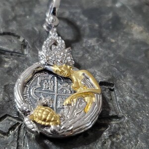 May include: Silver and gold pendant featuring a mermaid with a pearl crown and a gold turtle holding a silver coin with a cross design.