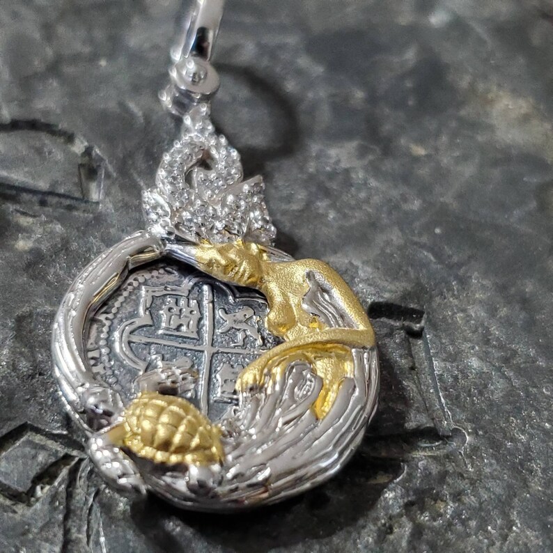 May include: Silver and gold pendant featuring a mermaid with a pearl crown and a gold turtle holding a silver coin with a cross design.