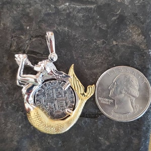 May include: A silver mermaid pendant with a gold tail, holding a silver coin with a cross design. The pendant is about the size of a UK 20 pence coin.