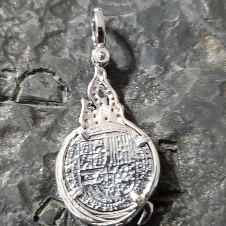 May include: Silver pendant featuring a detailed replica of a Spanish colonial coin with a decorative flame design on the bail.