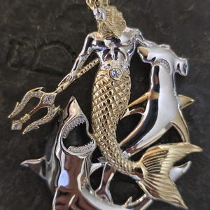 May include: A two-tone metal pendant featuring a mythical sea king with a trident, a crown, and a fish tail. The pendant is surrounded by two sharks. The king's torso is silver, and his tail and crown are gold. The sharks are silver.