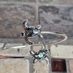 May include: A silver-toned bracelet featuring two detailed turtle charms. Each turtle has a textured shell with iridescent abalone-like inlays. The bracelet has a simple, open design, showcasing the turtle motifs as the focal point. The bracelet is a piece of jewelry.