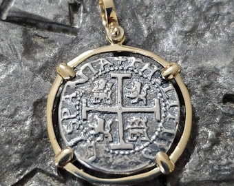 Puede incluir: Colgante de moneda de plata con bisel y fianza dorados. La moneda presenta un diseño de cruz con símbolos heráldicos. El colgante es probablemente de plata y oro, y es una pieza de joyería.