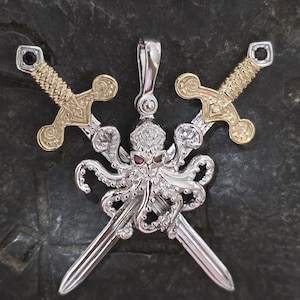 May include: A silver-toned pendant featuring a central octopus with red gemstone eyes, positioned over two crossed swords. The swords have gold-toned hilts with black gemstone accents at the pommels. A silver bail is attached to the top of the octopus.