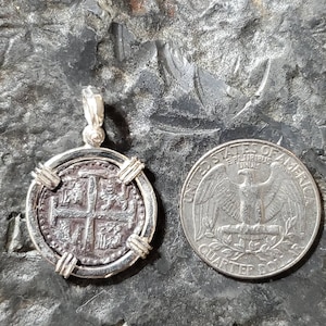 May include: A silver pendant featuring a replica of a Spanish colonial coin with a cross design. The coin is surrounded by a silver frame with four decorative accents. The pendant is shown next to a US quarter for size comparison.