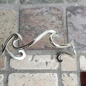 May include: A silver-colored, wave-shaped bracelet and a smaller matching piece. The bracelet curves in a flowing design, with a polished, reflective surface. The pieces are displayed on a textured, multi-colored stone surface.
