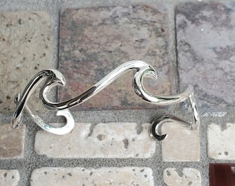 May include: A silver-colored, wave-shaped bracelet and a smaller matching piece. The bracelet curves in a flowing design, with a polished, reflective surface. The pieces are displayed on a textured, multi-colored stone surface.
