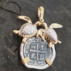 May include: A gold and silver pendant featuring two turtles flanking a silver coin. The coin has an intricate design with a cross in the center. The pendant has a gold bail for hanging.