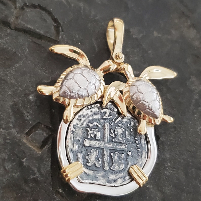 May include: A gold and silver pendant featuring two turtles flanking a silver coin. The coin has an intricate design with a cross in the center. The pendant has a gold bail for hanging.