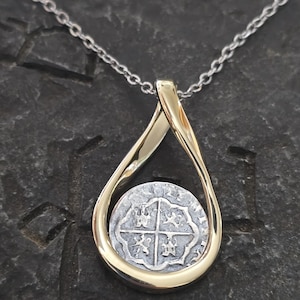 May include: A silver coin pendant necklace with a gold-colored teardrop-shaped bail. The coin has a cross design. The necklace chain is silver. The pendant is displayed against a dark, textured background.