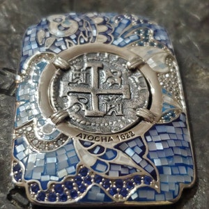 May include: A silver and blue mosaic pendant featuring a silver coin with a cross design and the text "ATOCHA 1622".