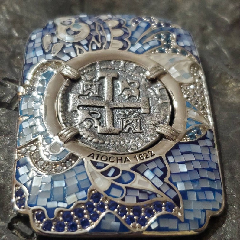 May include: A silver and blue mosaic pendant featuring a silver coin with a cross design and the text "ATOCHA 1622".