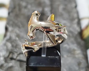 May include: A silver-colored hammerhead shark ring. The ring features a detailed design of the shark's head and body, with a wrap-around style. The shark's fin has a yellow accent. The ring is displayed on a black stand.