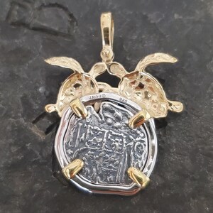 May include: A silver and gold pendant featuring a coin design. The pendant has a gold bail and accents, with two gold turtle figures embracing the coin. The coin has a detailed, textured surface. The pendant is marked with "J.Spirit" and "925".