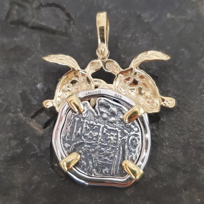 May include: A silver and gold pendant featuring a coin design. The pendant has a gold bail and accents, with two gold turtle figures embracing the coin. The coin has a detailed, textured surface. The pendant is marked with "J.Spirit" and "925".