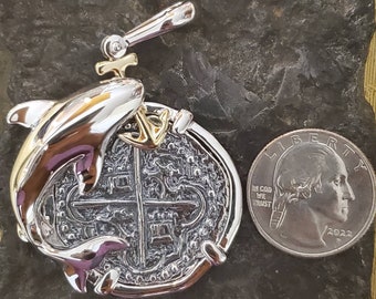 May include: A silver and gold pendant featuring a dolphin jumping over a replica of a Spanish eight reales coin. The coin has a cross design and the inscription "IN COD WE TRUST" is visible on the quarter dollar coin for size comparison.