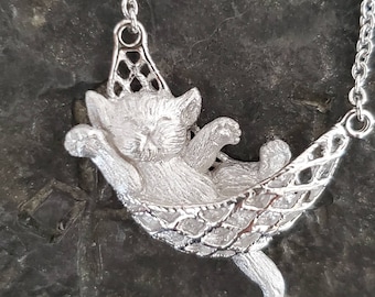 Op de afbeelding: Een zilveren hangerketting met een slapende kat in een hangmat. De kat is gedetailleerd met vacht en heeft een ontspannen uitdrukking. De hangmat is gemaakt van een delicaat, ingewikkeld ontwerp.