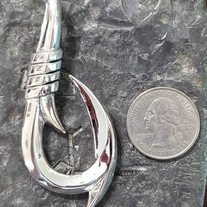 May include: A polished silver metal fish hook pendant. The hook is next to a U.S. quarter for size comparison. The hook has a detailed design with a wrapped section near the top and a sharp, curved point. The pendant is on a textured, dark gray surface.
