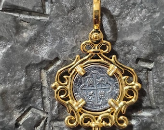 Op de afbeelding: Een gouden hanger met een zilveren munt met een kruis en vier symbolen. De munt is geplaatst in een decoratief gouden frame.