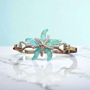 May include: A silver-toned bracelet featuring a large, iridescent blue-green starfish centerpiece. The starfish is adorned with a smaller, silver-toned starfish in the center. The bracelet has a hinged design.