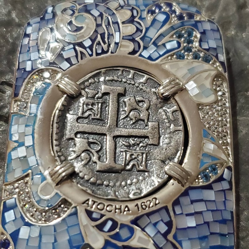 May include: A silver pendant with a blue and white mosaic design. The pendant features a replica of a Spanish silver coin with the inscription "ATOCHA 1622".