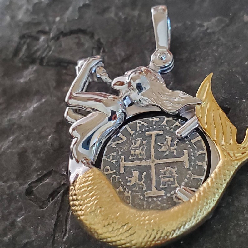 May include: A silver mermaid pendant with a gold tail holding a silver coin with a cross design. The coin has the text "PATRIA" and "LIBERTAD" around the edge.
