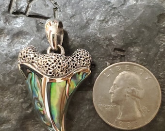 May include: A silver pendant in the shape of a shark tooth, featuring a colorful abalone shell inlay. The pendant has a textured, openwork design at the top and is shown next to a U.S. quarter for size comparison.