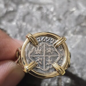 May include: A gold ring featuring a silver coin with the year 1622. The coin has a cross design and is set in a gold band with four gold bars holding the coin in place. The ring is a unique piece of jewelry.